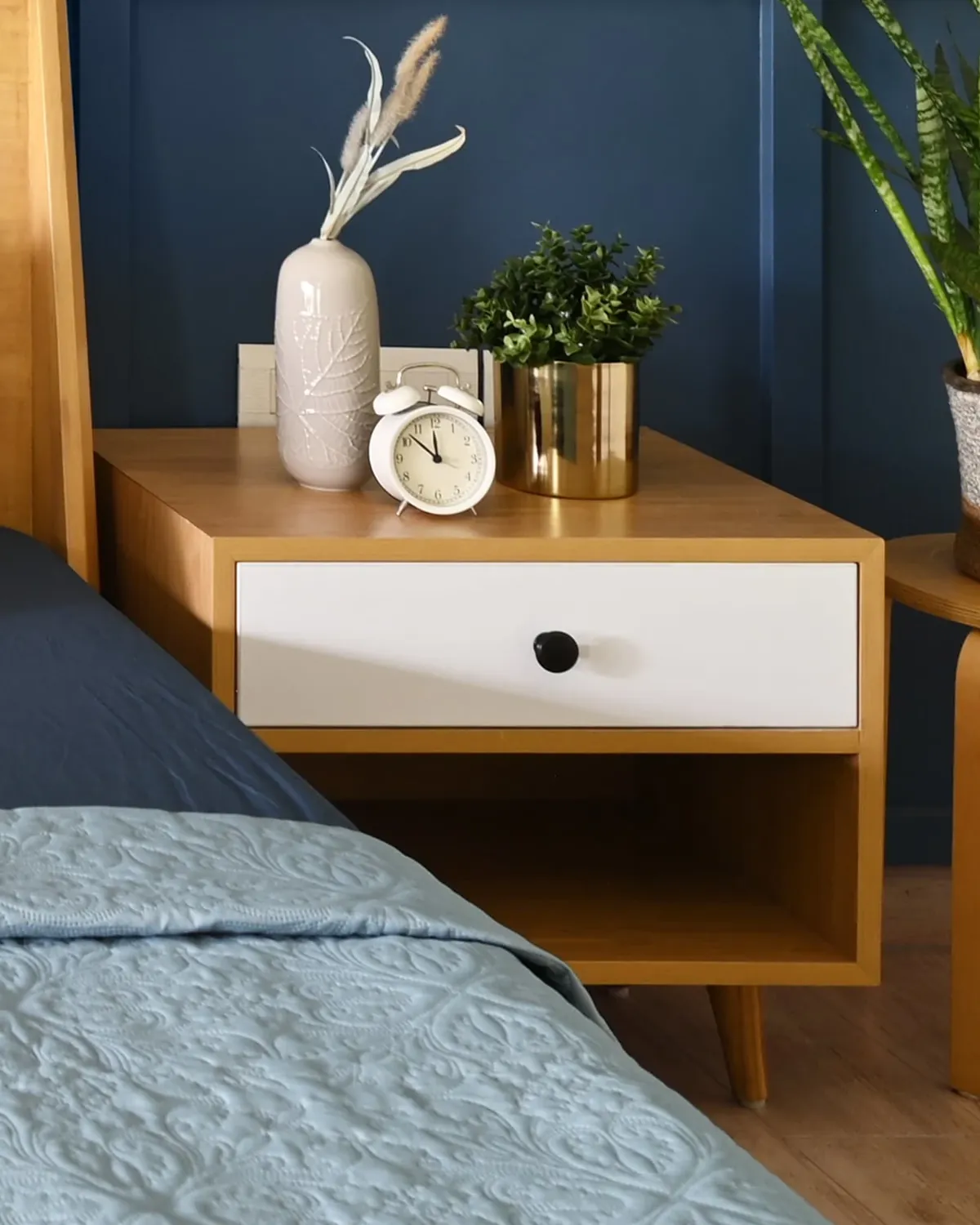 A teak bedside table with a white drawer over an open shelf, a clock and a vase on top.