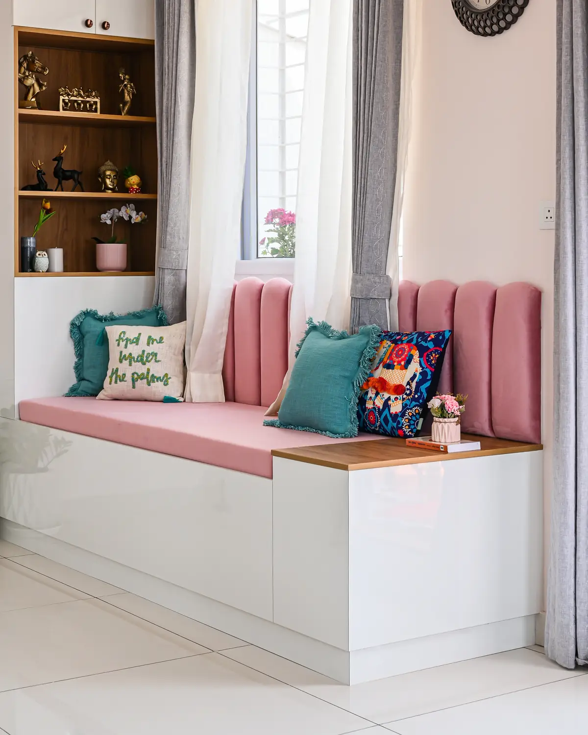 A diwan built into a window, a white base with drawers, a pink velvet seat and back, cushions on it.
