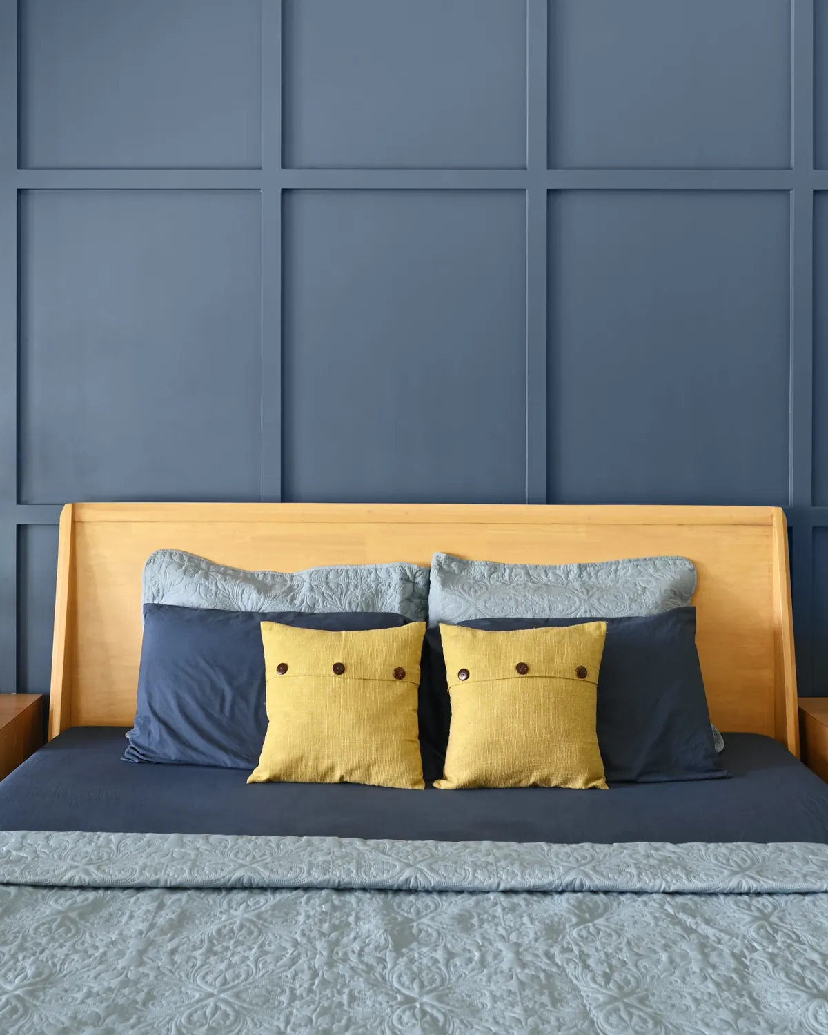 A teak king bed with a tall plain headboard and navy and yellow cushions, before a blue panelled wall.