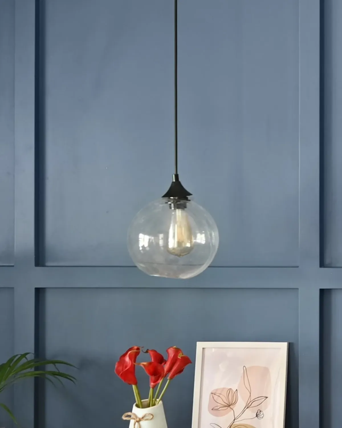 A clear glass globe pendant with a filament bulb, before a blue panelled wall.