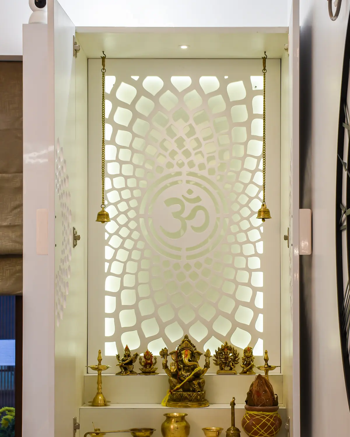 A white pooja unit, its doors open on a backlit jaali panel cut with Om, brass idols on the shelf.