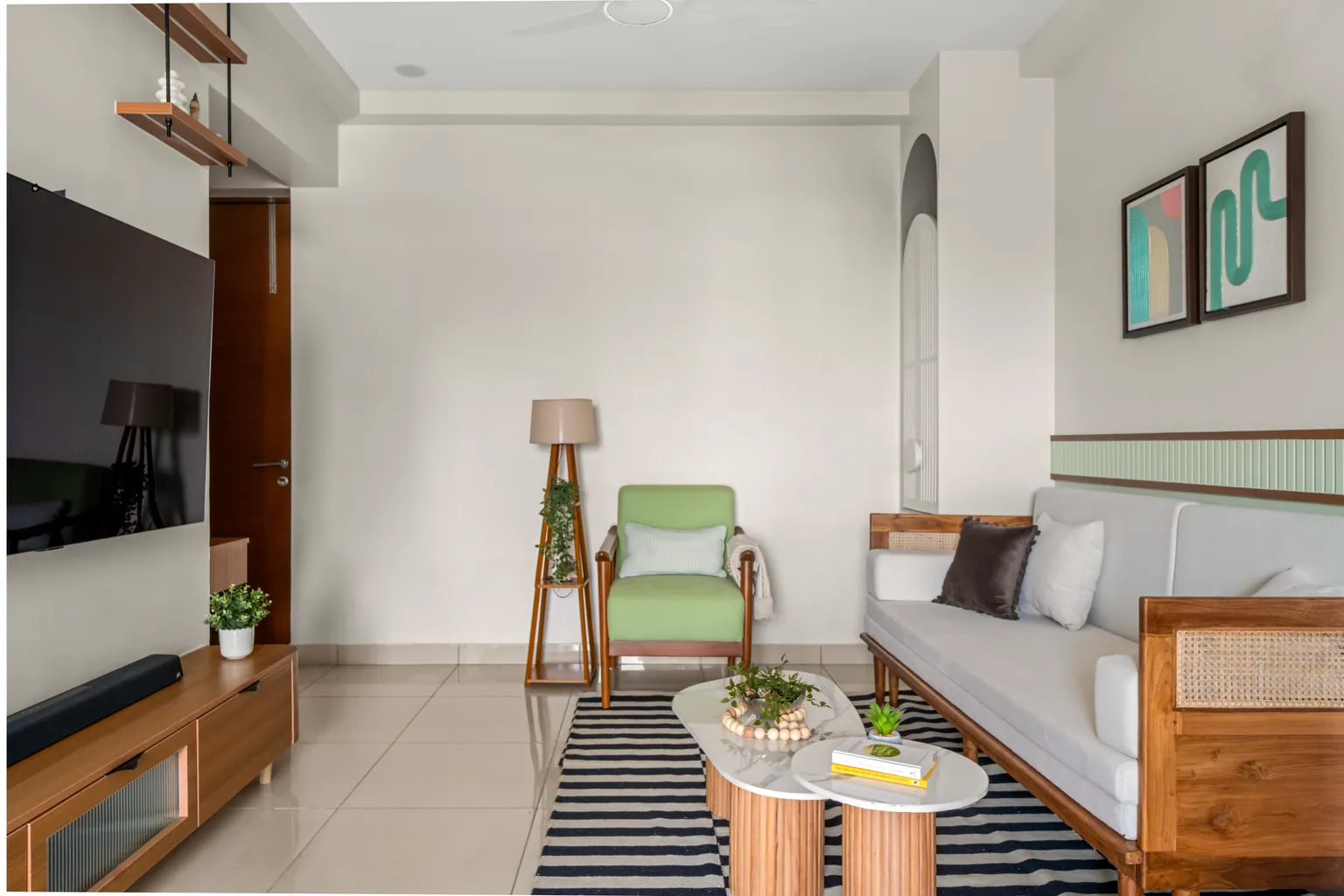A living room with a teak sofa with cane sides, a sage green armchair, a teak floor lamp, marble-topped tables on a striped rug and teak shelves hung above the TV.