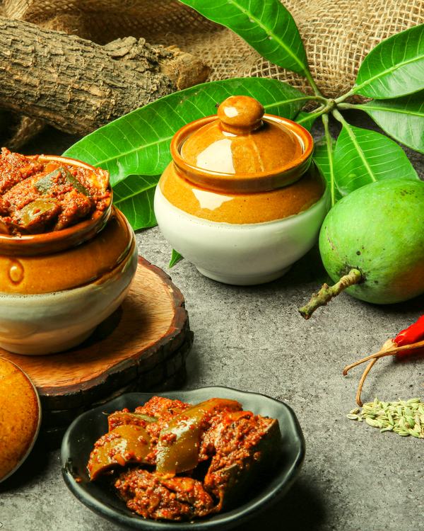 Mango pickle in ceramic jars, raw mangoes and red chillies beside them.