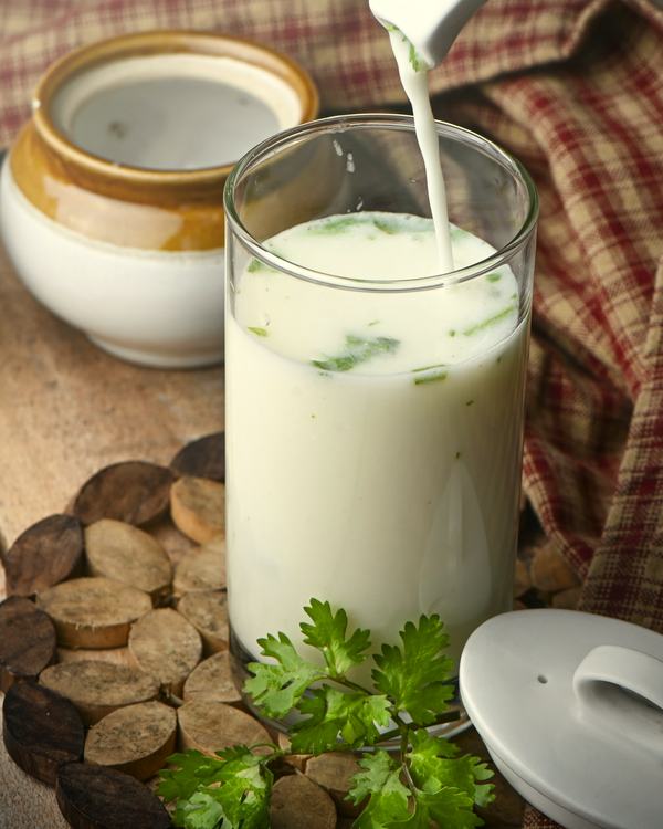 Majjiga poured into a tall glass, coriander and a lidded pot beside it.