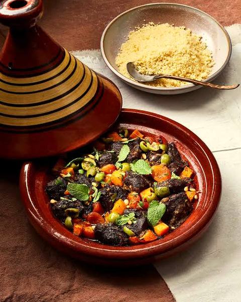A clay pot with its tall lid lifted off a stew of meat and vegetables, a bowl of grain beside it.