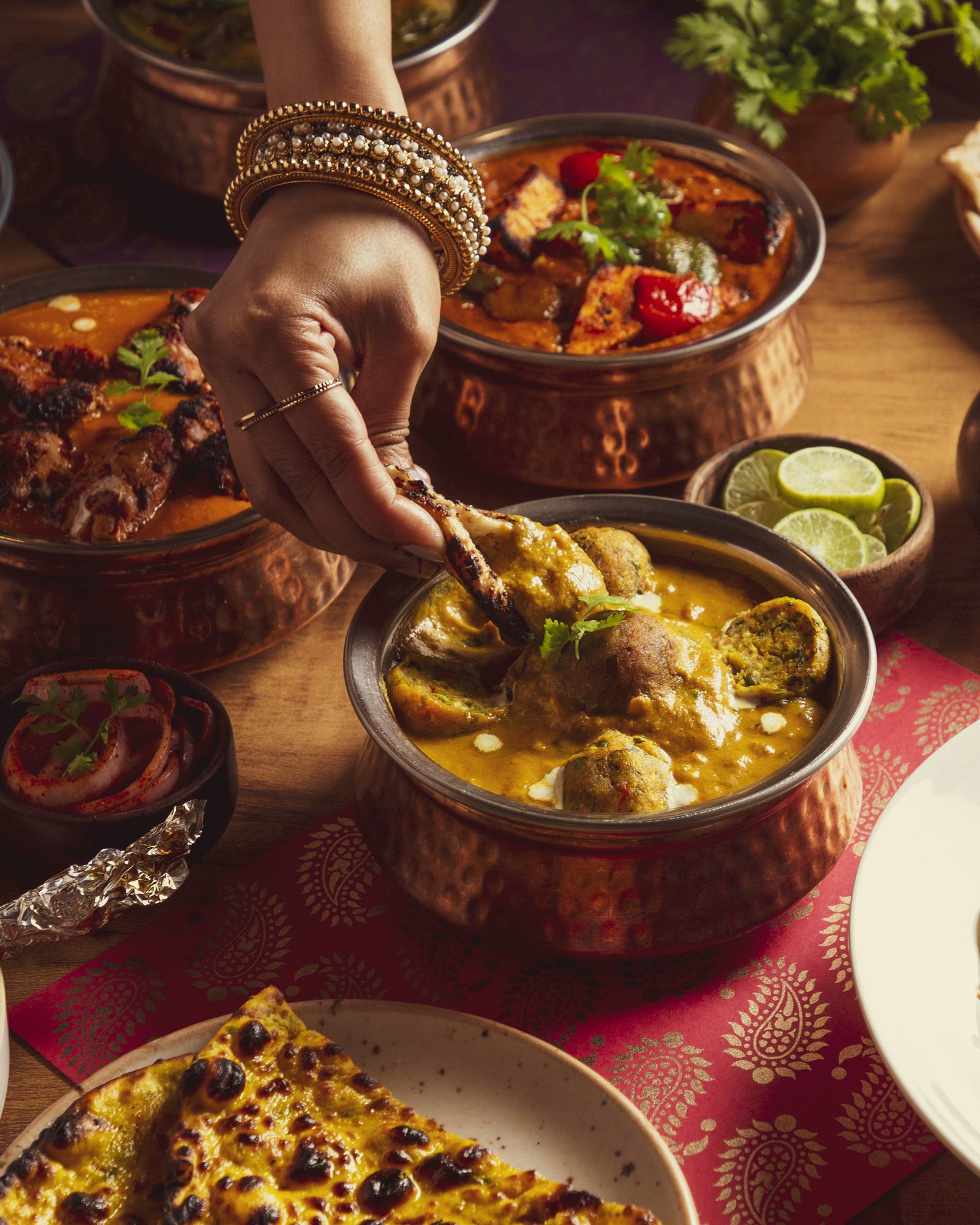 A table of curries in copper handis, naan and noodles, a hand with bangles reaching in.