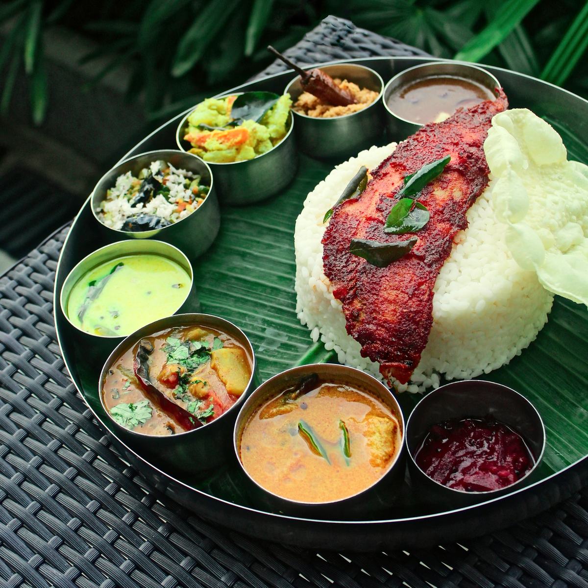 A lunch thali from above: rice and fried fish on a banana leaf, small steel bowls of curry around it.