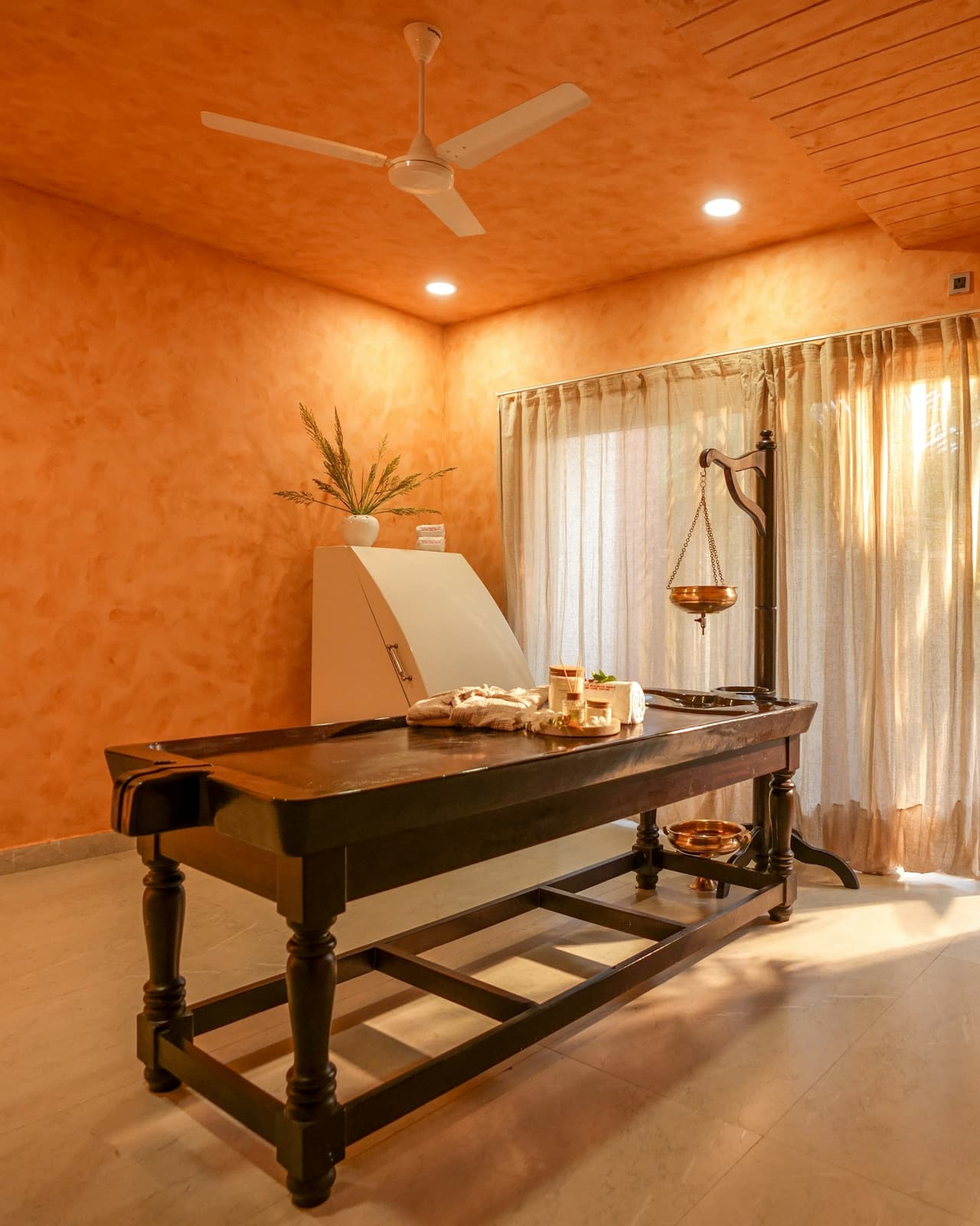 The spa room: a wooden oil table, a brass dhara pot on its stand, curtains over a bright door.